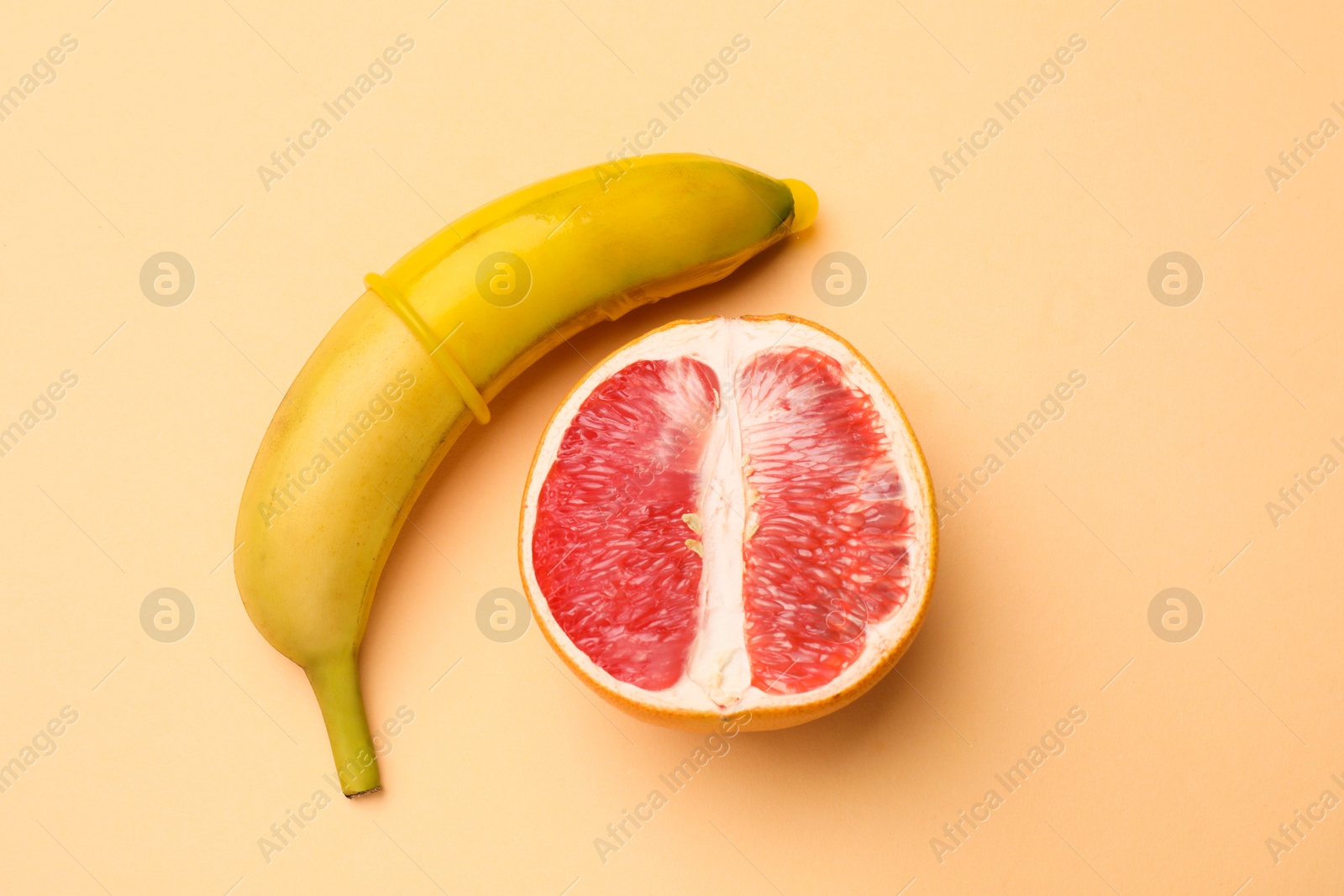 Photo of Sex education. Banana with condom and half of grapefruit on beige background, flat lay