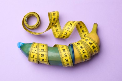 Photo of Sex education. Banana with condom and measuring tape on violet background, top view