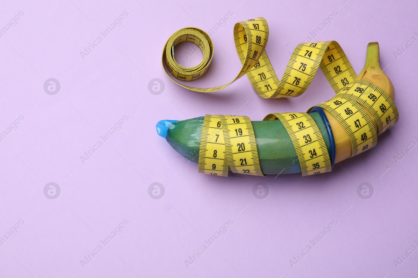Photo of Sex education. Banana with condom and measuring tape on violet background, top view. Space for text