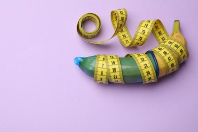 Photo of Sex education. Banana with condom and measuring tape on violet background, top view. Space for text