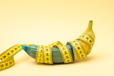 Photo of Sex education. Banana with condom and measuring tape on beige background