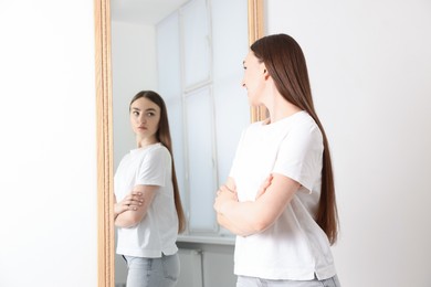 Image of Young woman looking at serious reflection of herself in mirror indoors. Mental health