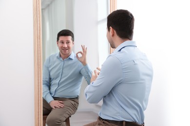 Image of Man sitting in front of mirror where his reflection showing Ok gesture indoors. Mental health