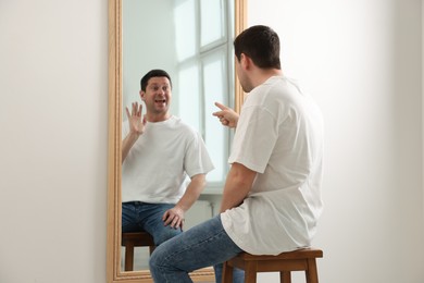 Image of Man pointing seriously at his cheerful reflection in mirror indoors. Mental health