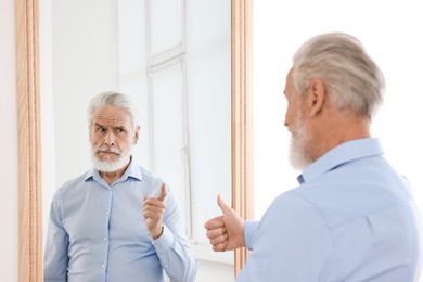 Image of Senior man showing thumbs up in front of mirror while his reflection demonstrating serious face expression indoors. Mental health