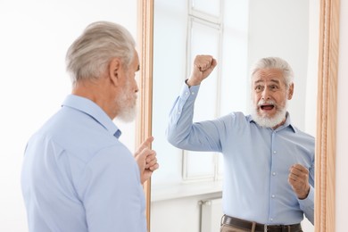 Image of Serious senior man looking at happy reflection of himself in mirror indoors. Mental health