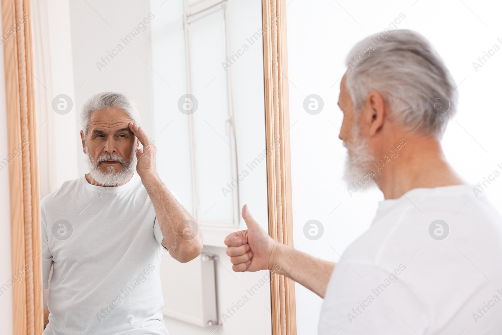 Image of Senior man showing thumbs up in front of mirror while his reflection demonstrating thoughtful face expression indoors. Mental health