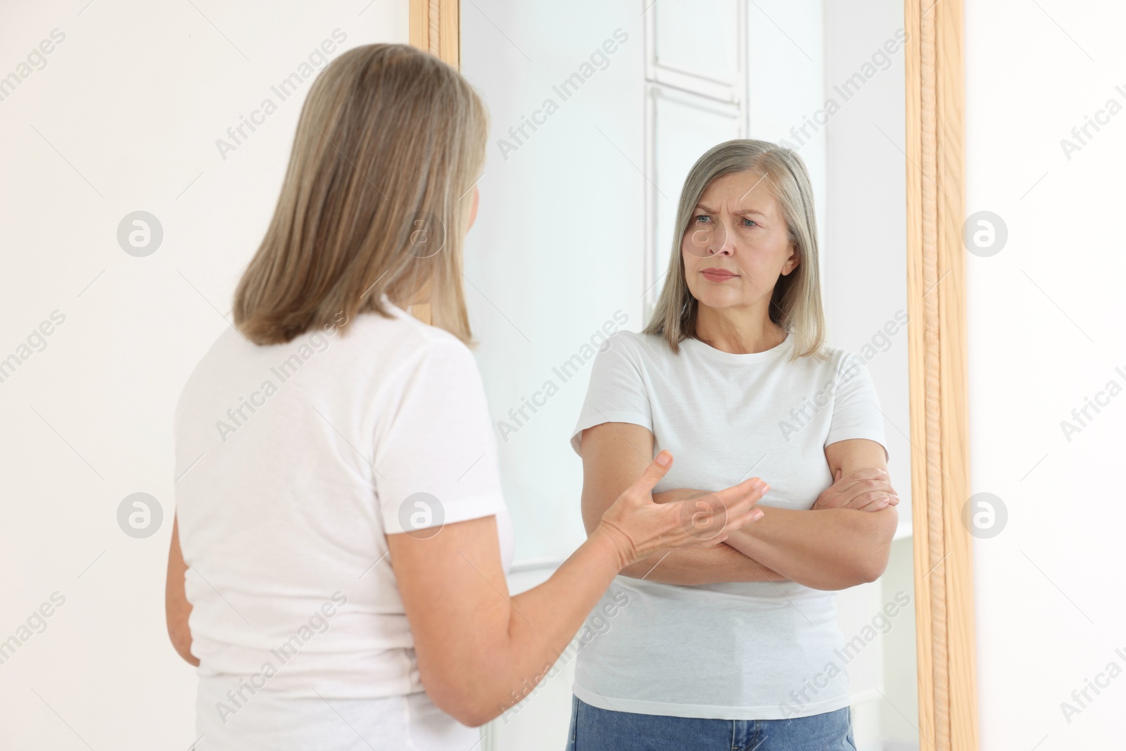 Image of Senior woman looking at serious reflection of herself in mirror indoors. Mental health