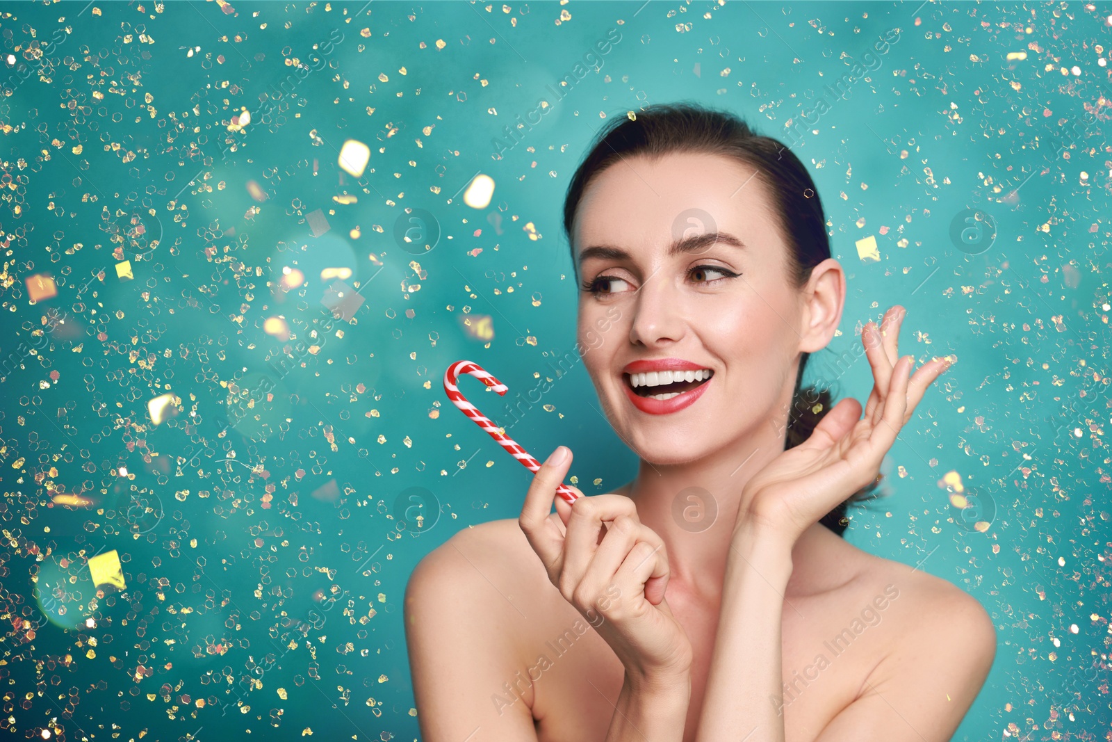 Smiling woman with perfect makeup holding Christmas candy cane against turquoise background with golden lights Image of Smiling woman with perfect makeup holding Christmas candy cane against turquoise background with golden lights