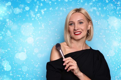 Image of Smiling woman holding makeup brush on light blue background with bokeh lights