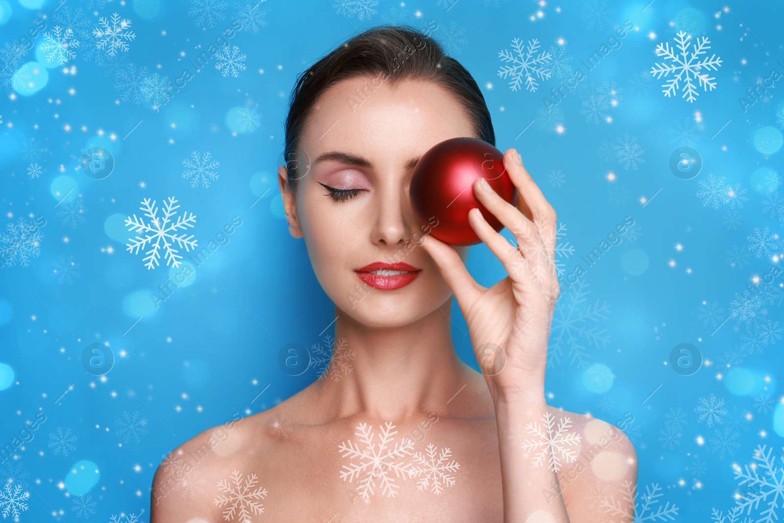 Woman with perfect makeup holding Christmas bauble on light blue background with snowflakes Image of Woman with perfect makeup holding Christmas bauble on light blue background with snowflakes