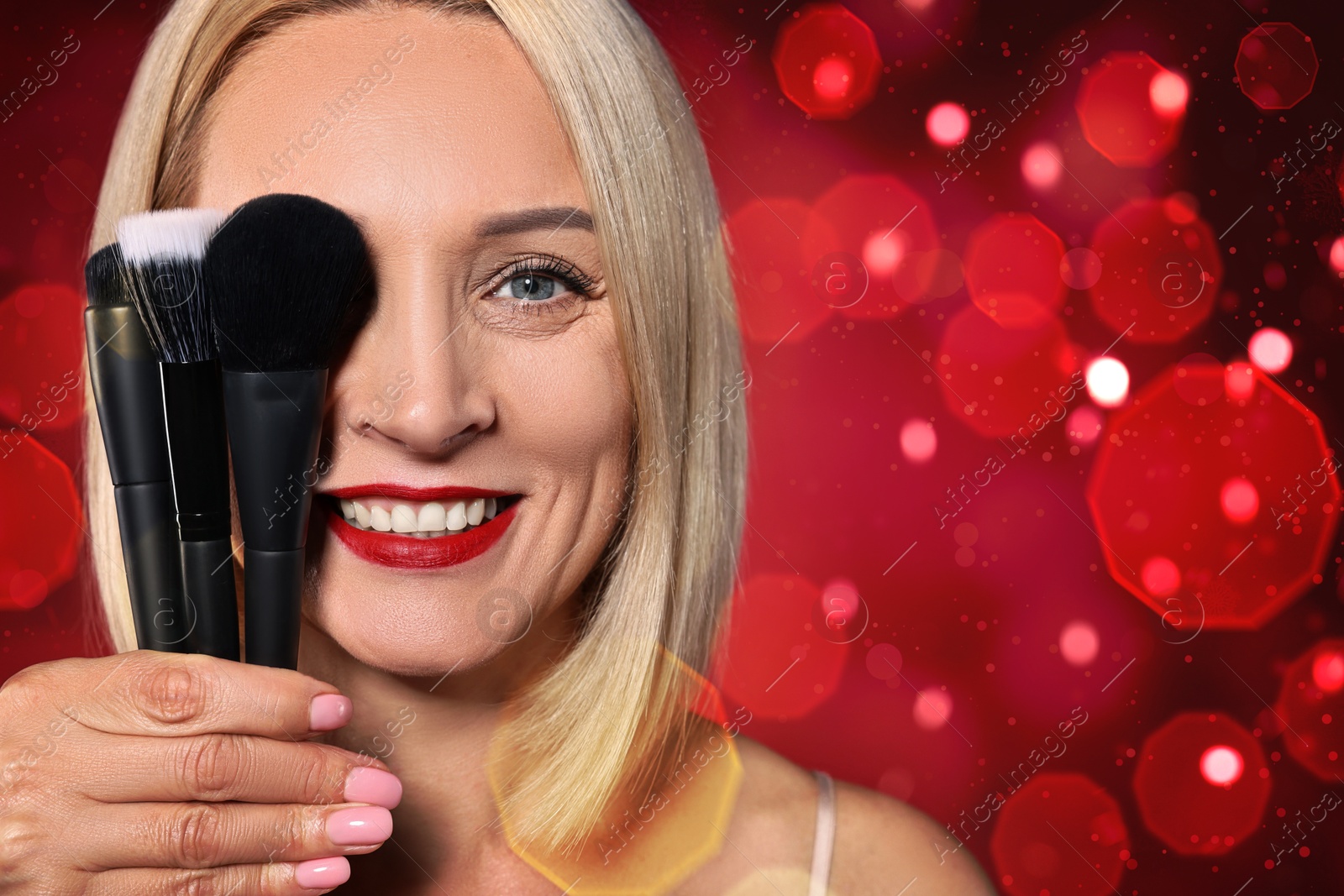 Smiling woman holding different makeup brushes on red background with bokeh lights Image of Smiling woman holding different makeup brushes on red background with bokeh lights