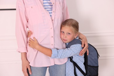 Photo of Little girl with backpack afraid to leave without mother and hugging her near white wall at home. Separation anxiety