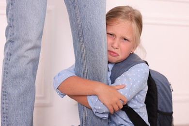 Photo of Little girl with backpack afraid to leave without mother and hugging her near white wall at home. Separation anxiety
