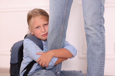 Photo of Little girl with backpack afraid to leave without mother and hugging her near white wall at home. Separation anxiety
