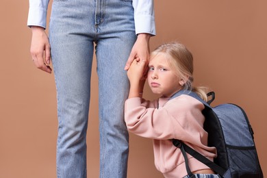 Photo of Little girl with backpack afraid to leave without mother and holding hands with her on brown background. Separation anxiety