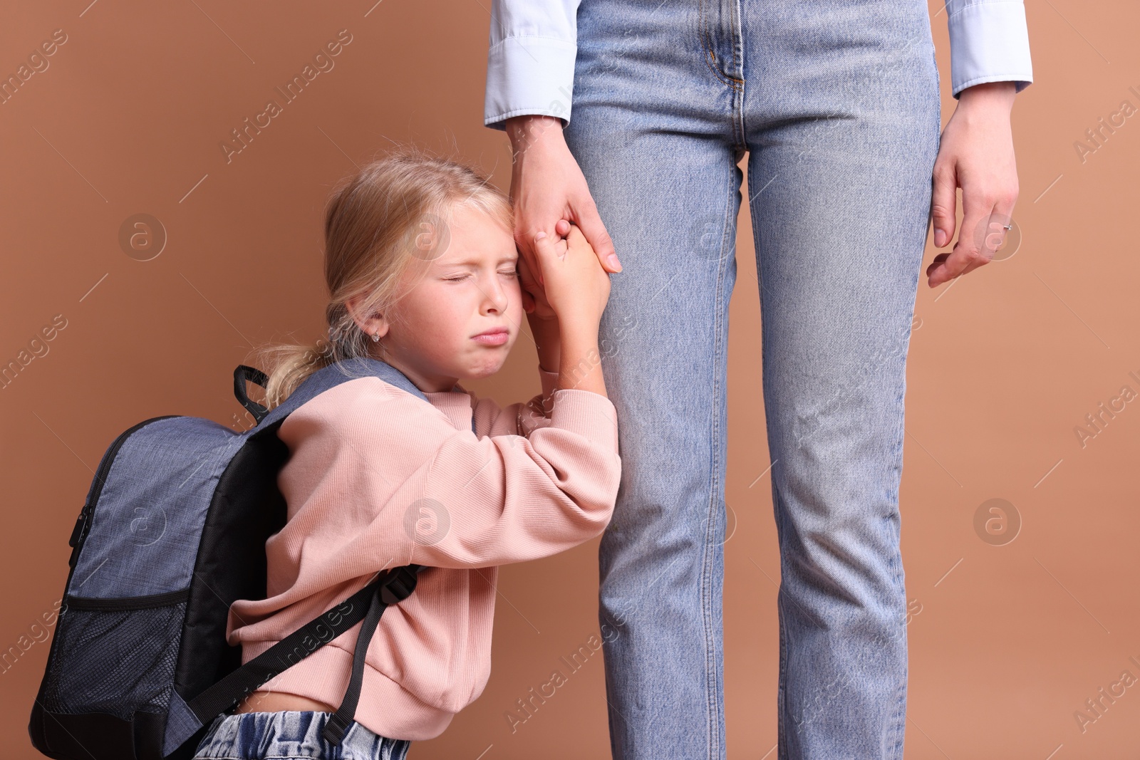 Photo of Little girl with backpack afraid to leave without mother and holding hands with her on brown background. Separation anxiety