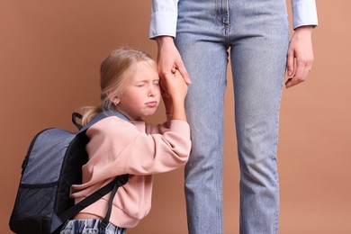 Photo of Little girl with backpack afraid to leave without mother and holding hands with her on brown background. Separation anxiety