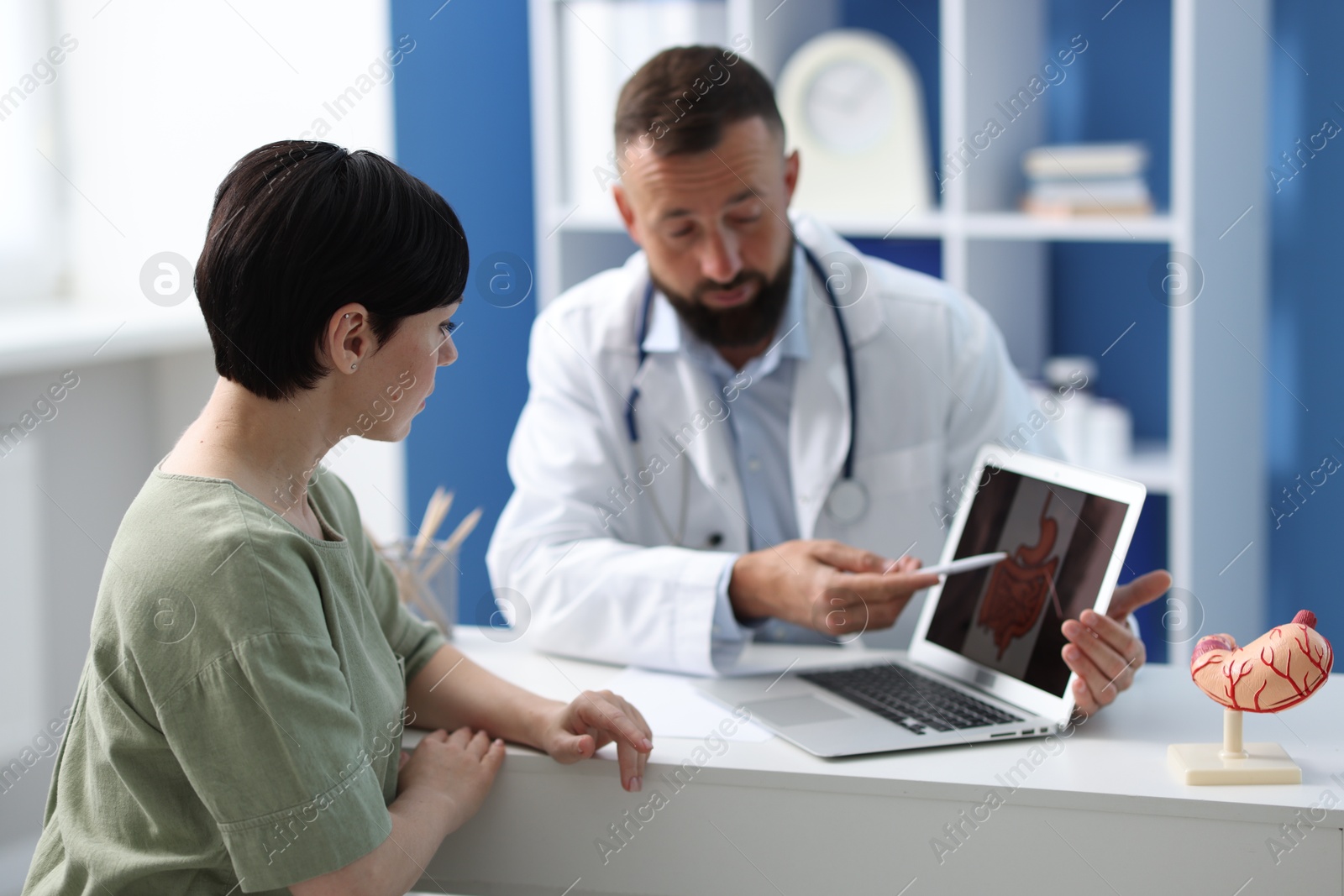 Photo of Gastroenterologist showing screen with illustration of large intestine to patient at table in clinic, selective focus