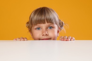 Photo of Curious little girl peeking over table against orange background