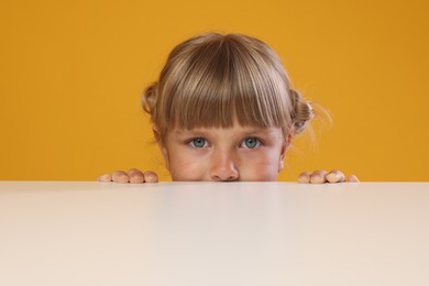 Photo of Curious little girl peeking over table against orange background