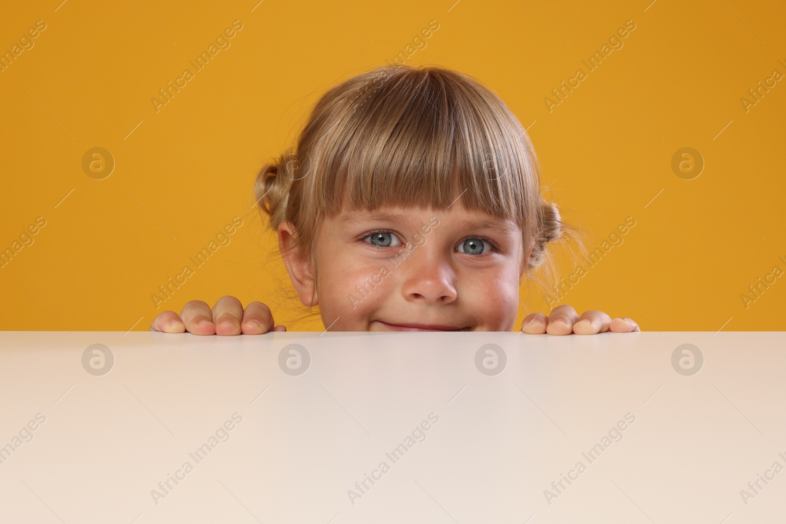 Photo of Curious little girl peeking over table against orange background
