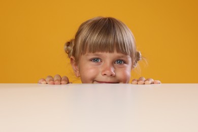 Photo of Curious little girl peeking over table against orange background