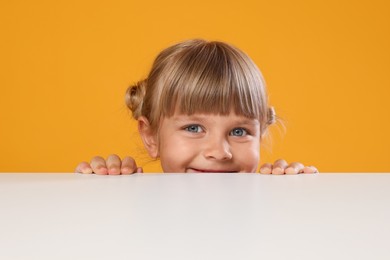 Photo of Curious little girl peeking over table against orange background