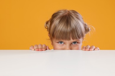 Photo of Curious little girl peeking over table against orange background