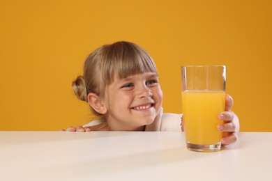 Photo of Cute little girl reaching for glass of juice at white table against orange background