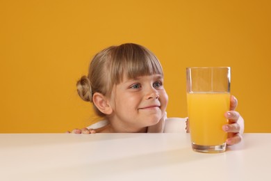Photo of Cute little girl reaching for glass of juice at white table against orange background