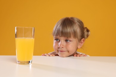 Photo of Cute little girl looking at glass of juice while peeking over table against orange background