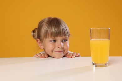 Photo of Cute little girl looking at glass of juice while peeking over table against orange background