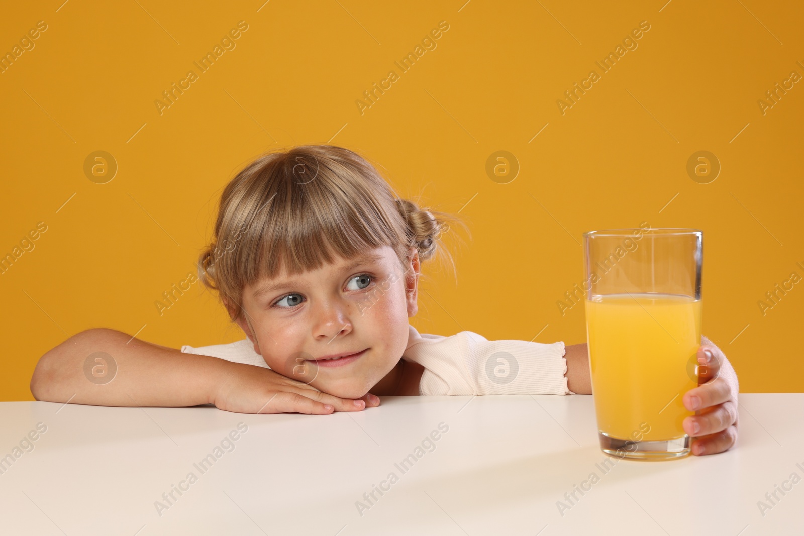 Photo of Cute little girl reaching for glass of juice at white table against orange background