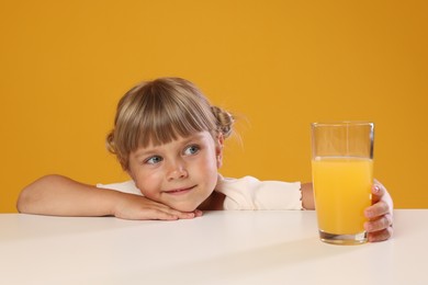 Photo of Cute little girl reaching for glass of juice at white table against orange background