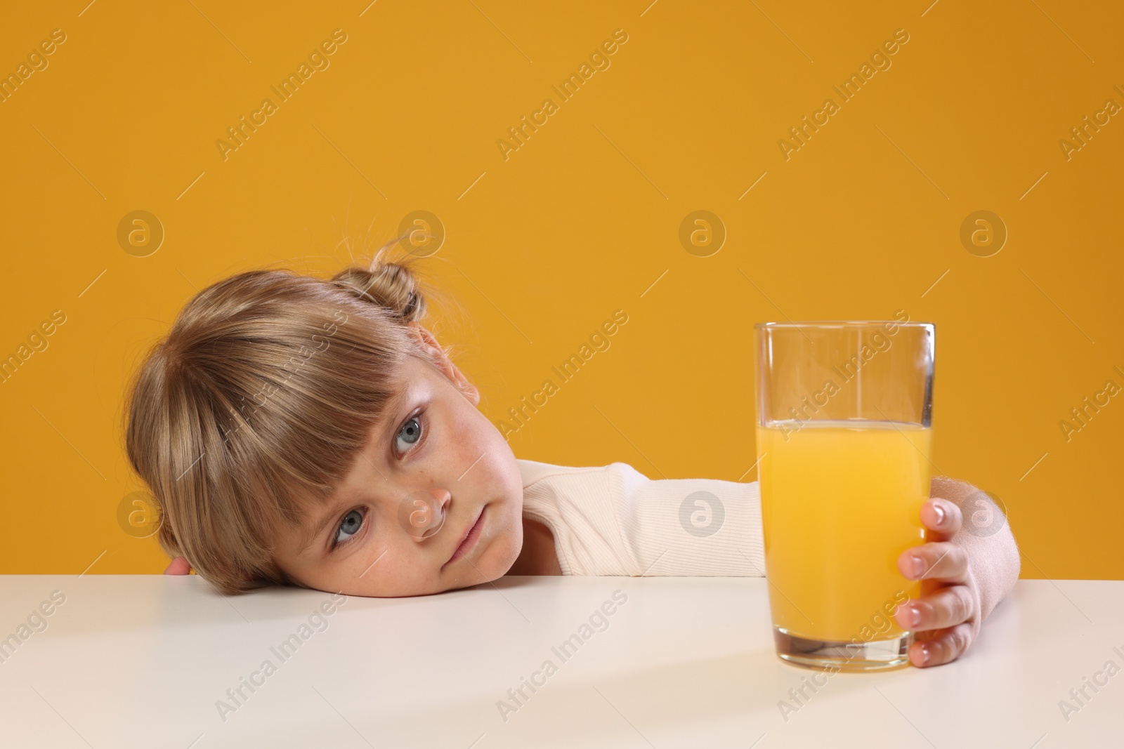 Photo of Cute little girl reaching for glass of juice at white table against orange background