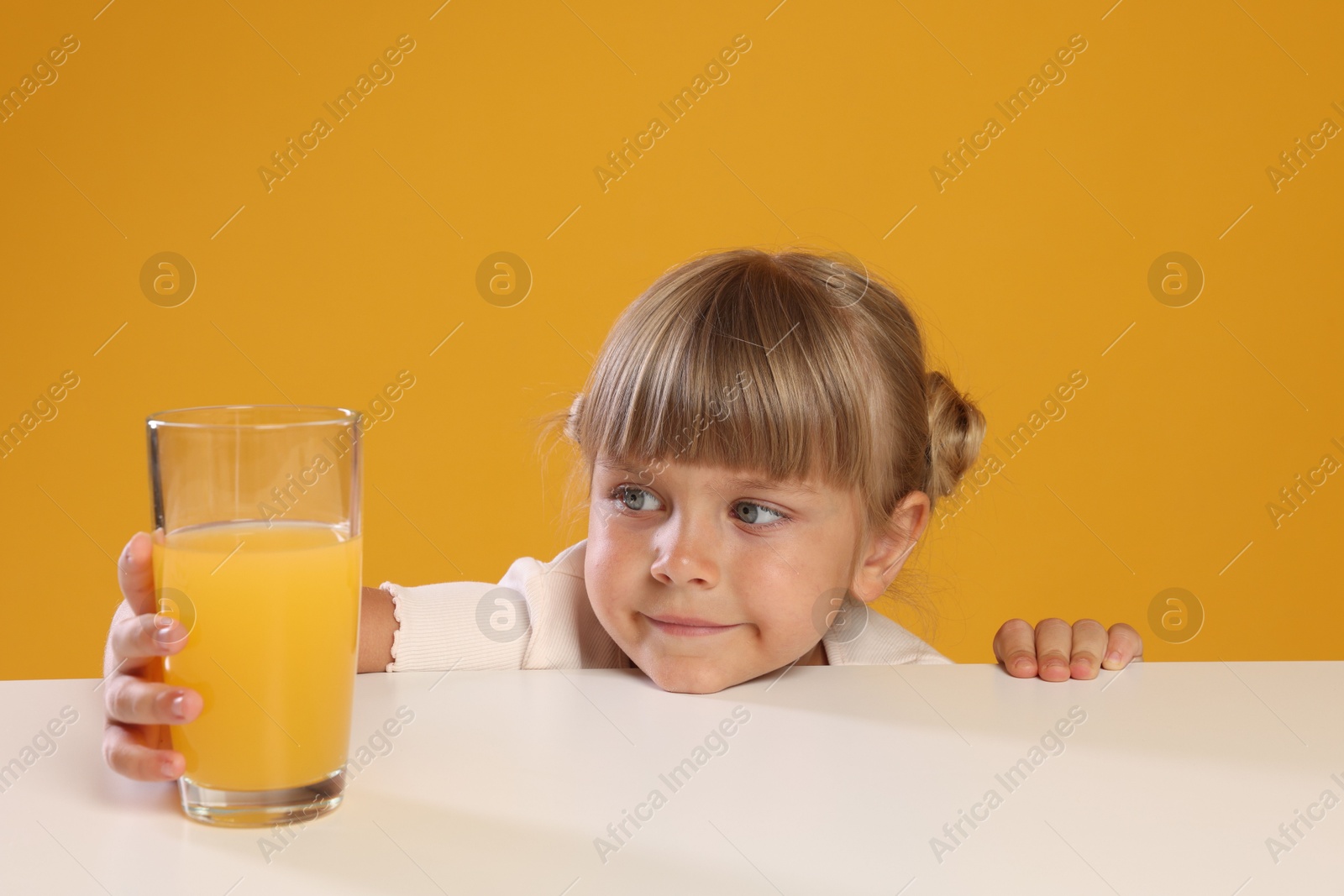 Photo of Cute little girl reaching for glass of juice at white table against orange background