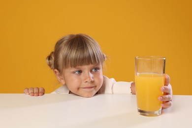 Photo of Cute little girl reaching for glass of juice at white table against orange background