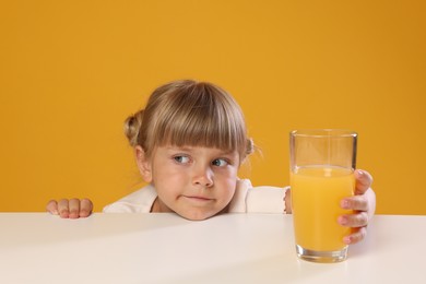 Photo of Cute little girl reaching for glass of juice at white table against orange background
