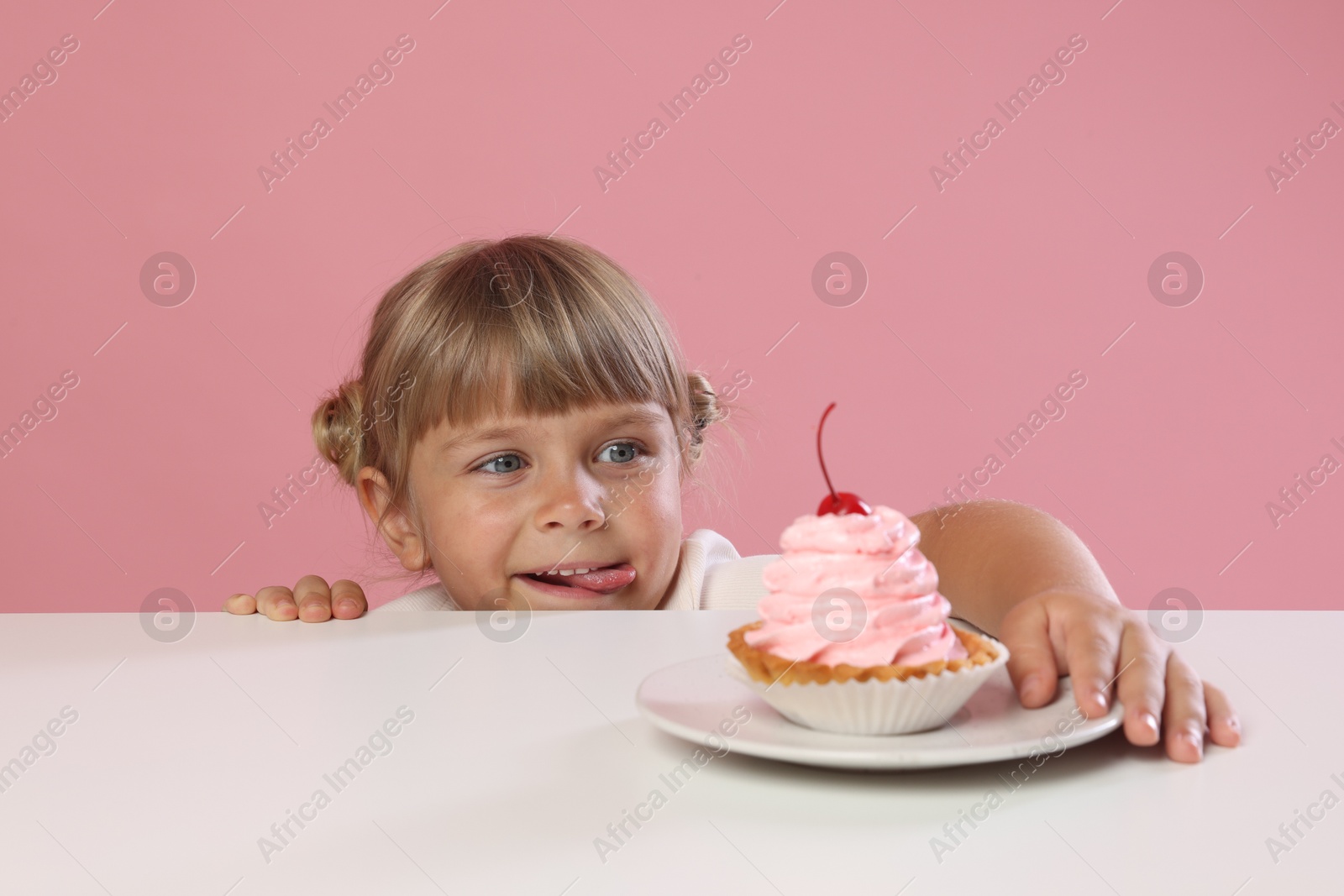 Photo of Cute little girl reaching for tasty dessert at white table against pink background