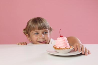 Photo of Cute little girl reaching for tasty dessert at white table against pink background