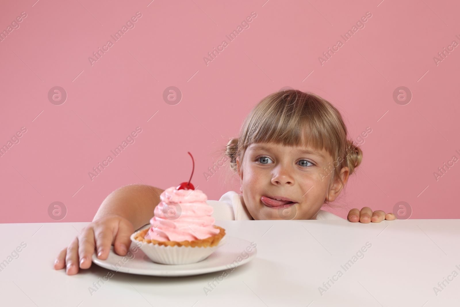 Photo of Cute little girl reaching for tasty dessert at white table against pink background