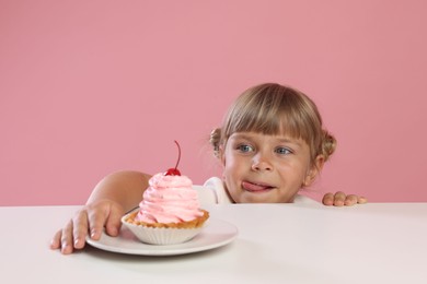 Photo of Cute little girl reaching for tasty dessert at white table against pink background