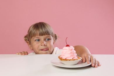 Photo of Cute little girl reaching for tasty dessert at white table against pink background