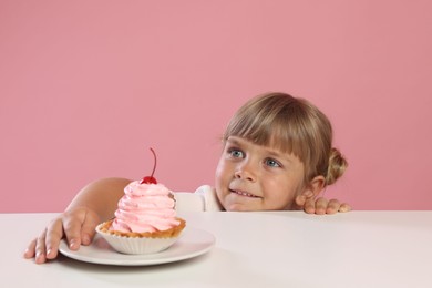Photo of Cute little girl reaching for tasty dessert at white table against pink background