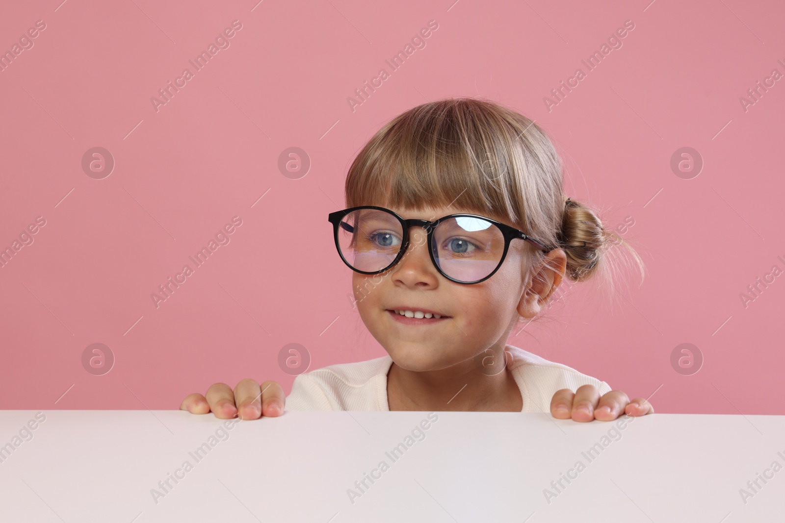 Photo of Curious little girl in glasses peeking over table against pink background