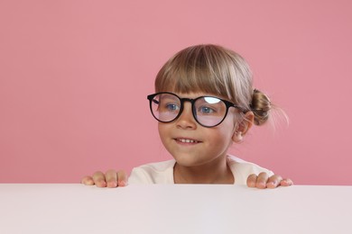 Photo of Curious little girl in glasses peeking over table against pink background