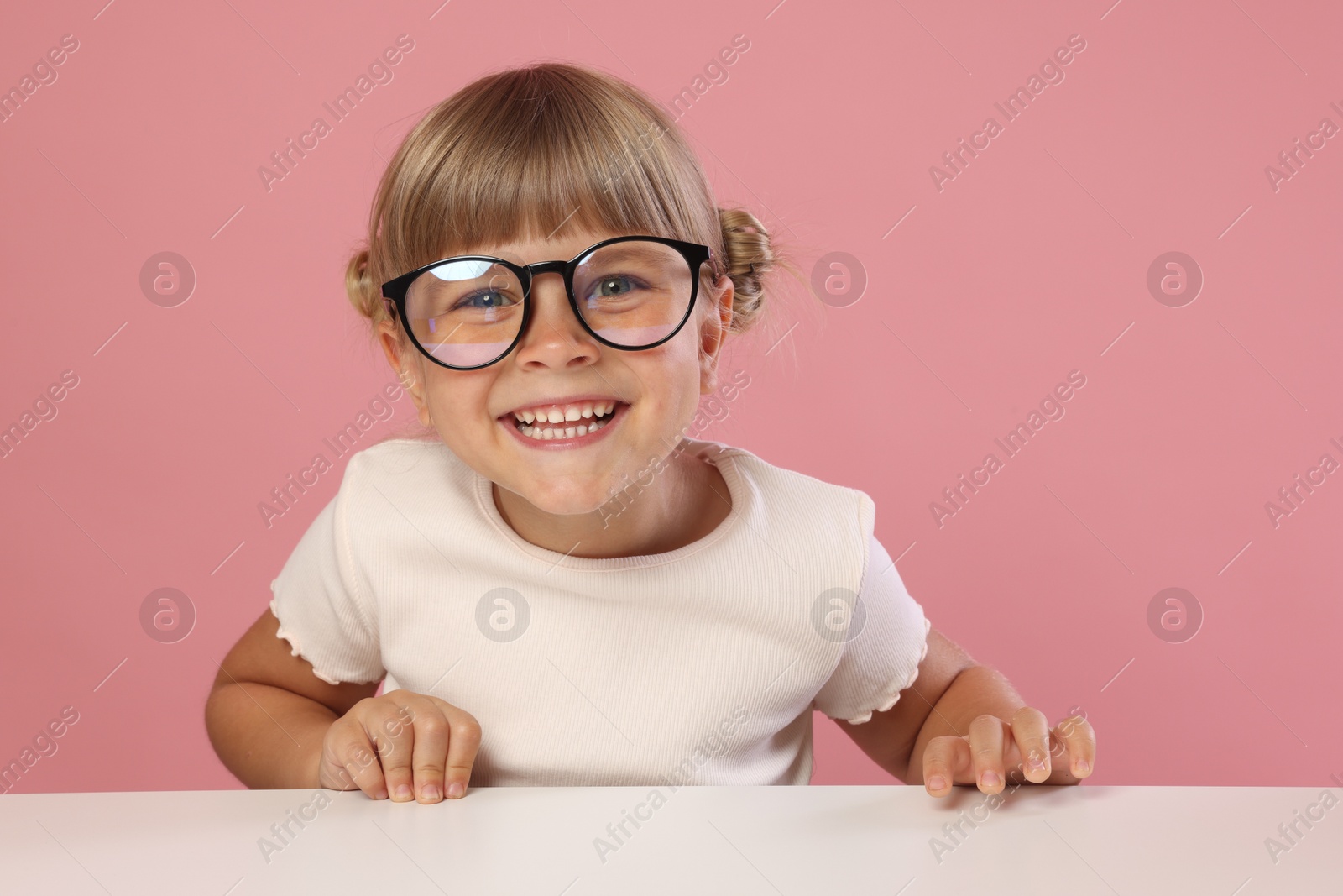 Photo of Curious little girl in glasses peeking over table against pink background