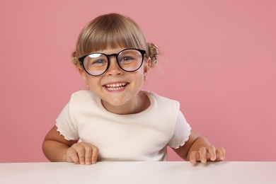 Photo of Curious little girl in glasses peeking over table against pink background