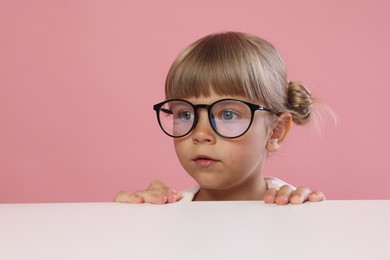 Photo of Curious little girl in glasses peeking over table against pink background, space for text